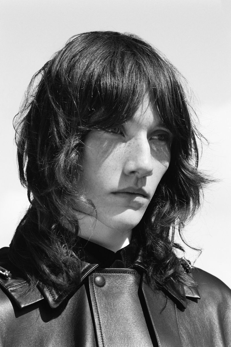 Monochrome portrait of a person with dark wavy hair wearing a black leather jacket against a bright sky, captured in natural light.