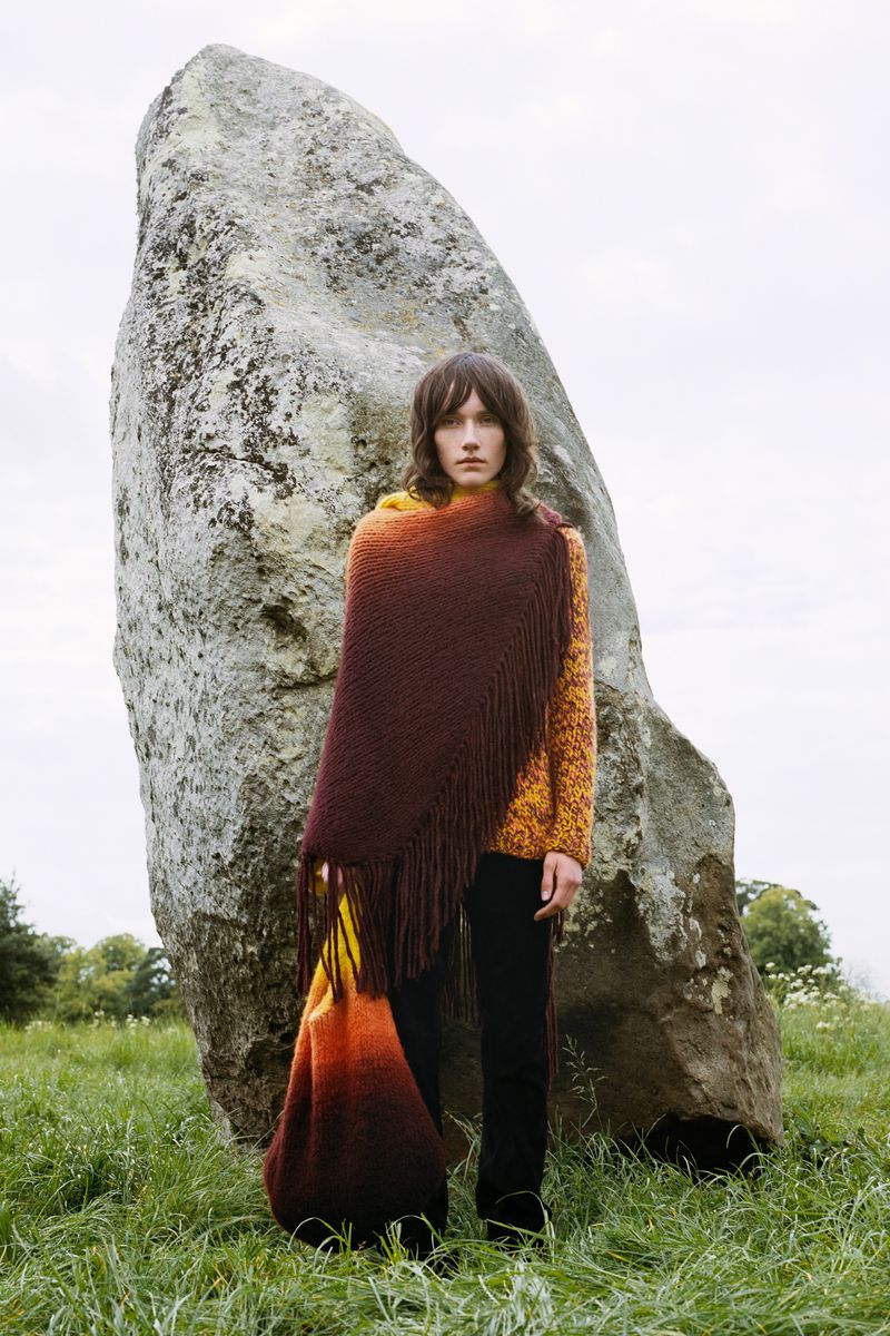 Woman stands in a field wearing brown shawl and yellow top, posing next to large rock formation in a rural outdoor scene.
