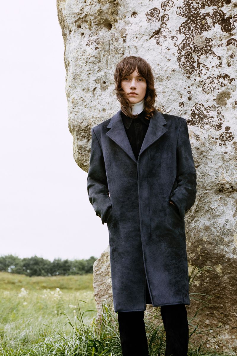 An adult male model with shoulder-length brown hair poses next to a large stone structure in a grassy field wearing a long blue overcoat.