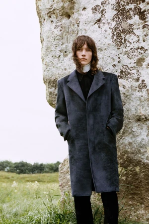 An adult male model with shoulder-length brown hair poses next to a large stone structure in a grassy field wearing a long blue overcoat.