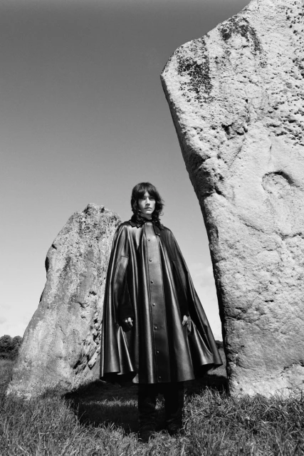 Monochrome portrait of person in black cape between standing stones in rural setting under blue sky. Fashion or editorial photography.