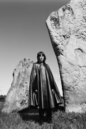 Monochrome portrait of person in black cape between standing stones in rural setting under blue sky. Fashion or editorial photography.