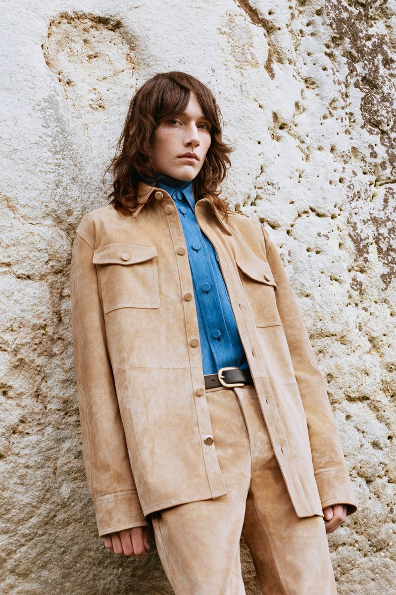 A person wearing a tan suede jacket and pants stands against a textured rock wall in a fashion editorial style full shot.