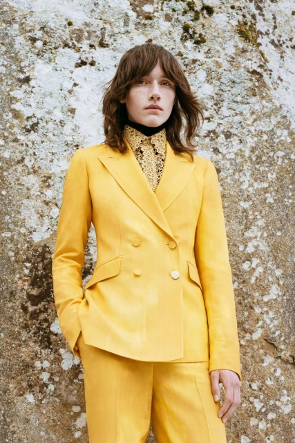 Woman in a yellow double-breasted suit, stands in front of a textured stone wall, looking directly at the camera in a fashion shot.