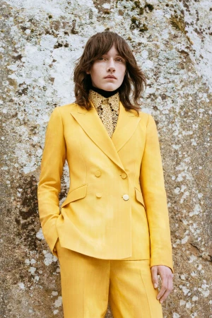 Woman in a yellow double-breasted suit, stands in front of a textured stone wall, looking directly at the camera in a fashion shot.
