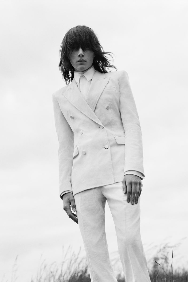 Monochrome portrait of a person in a light-colored double-breasted suit, standing in a field with a cloudy sky backdrop.