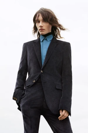 A person stands against a bright backdrop, wearing a corduroy suit jacket, matching pants, a blue shirt, and a black turtleneck.