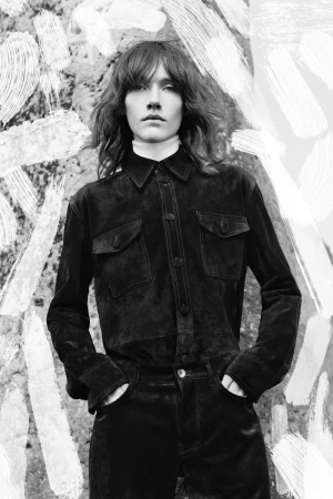 Model wearing a black suede button-up shirt and trousers with hands in pockets framed by painterly white brushstrokes.