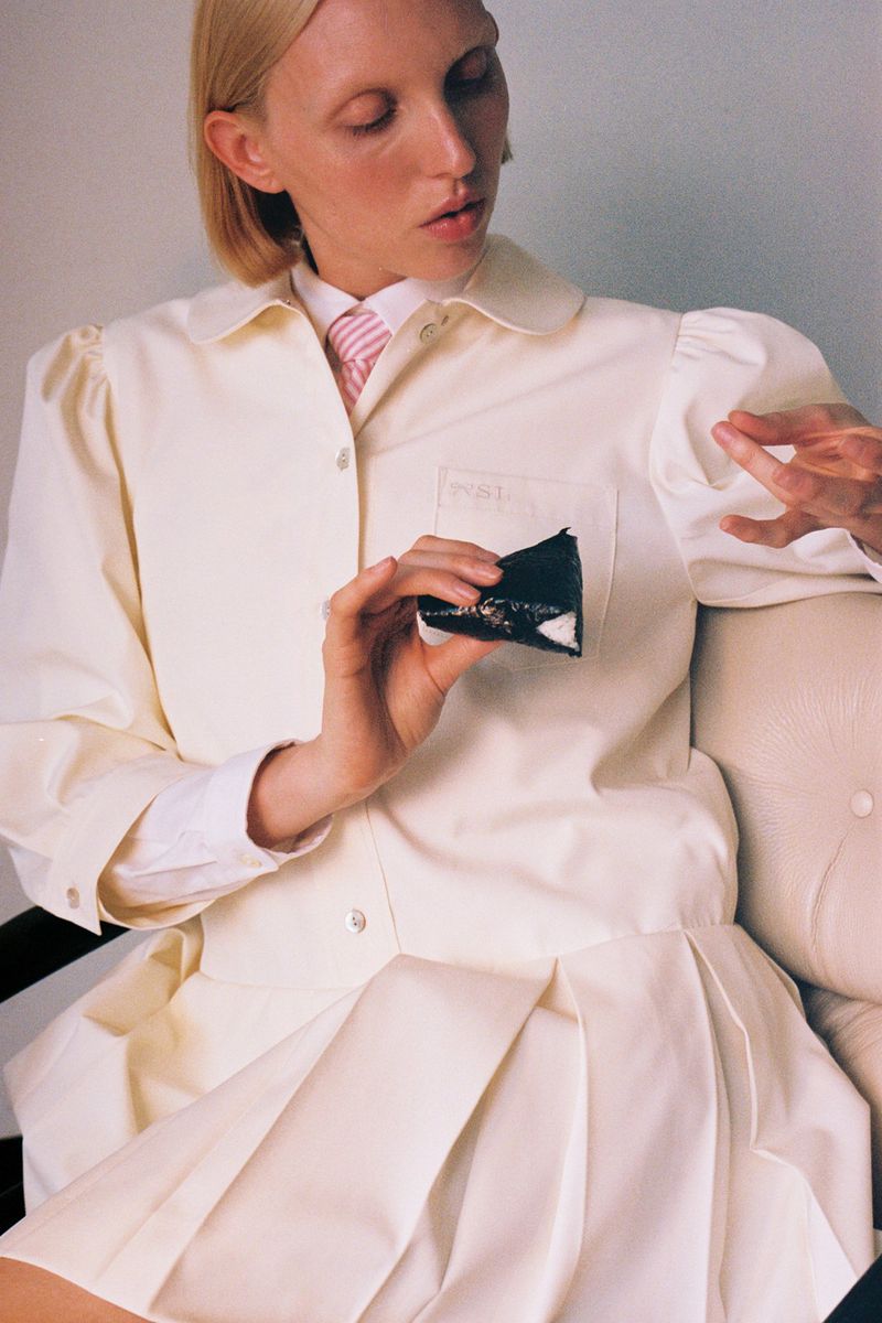 Woman in a cream-colored lab coat holds a black object while seated. She has pale skin, short blonde hair, and a pink tie.