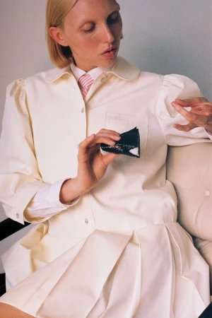 Woman in a cream-colored lab coat holds a black object while seated. She has pale skin, short blonde hair, and a pink tie.