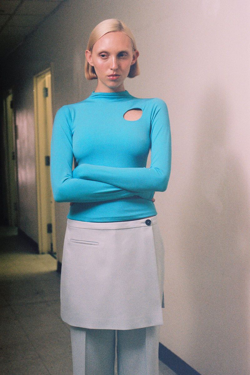 Woman in a turquoise long-sleeved shirt and wrap skirt poses in a hallway; modern fashion editorial with clean lines and muted colors.
