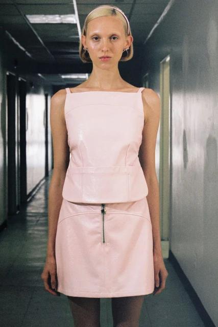 Woman stands in a hallway wearing a pink top and skirt outfit. She is looking directly at the camera in a studio shot.