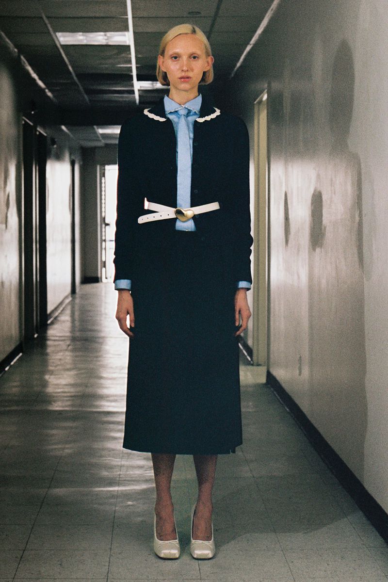 A slender blonde woman stands in a formal navy skirt suit with white heels in an indoor hallway.