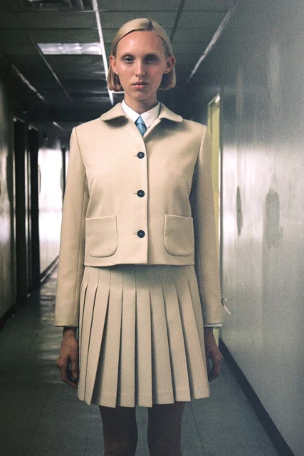 Fashion shot of a fair-skinned, blonde adult female in a beige, skirt suit standing in a hallway with light reflecting off the walls.