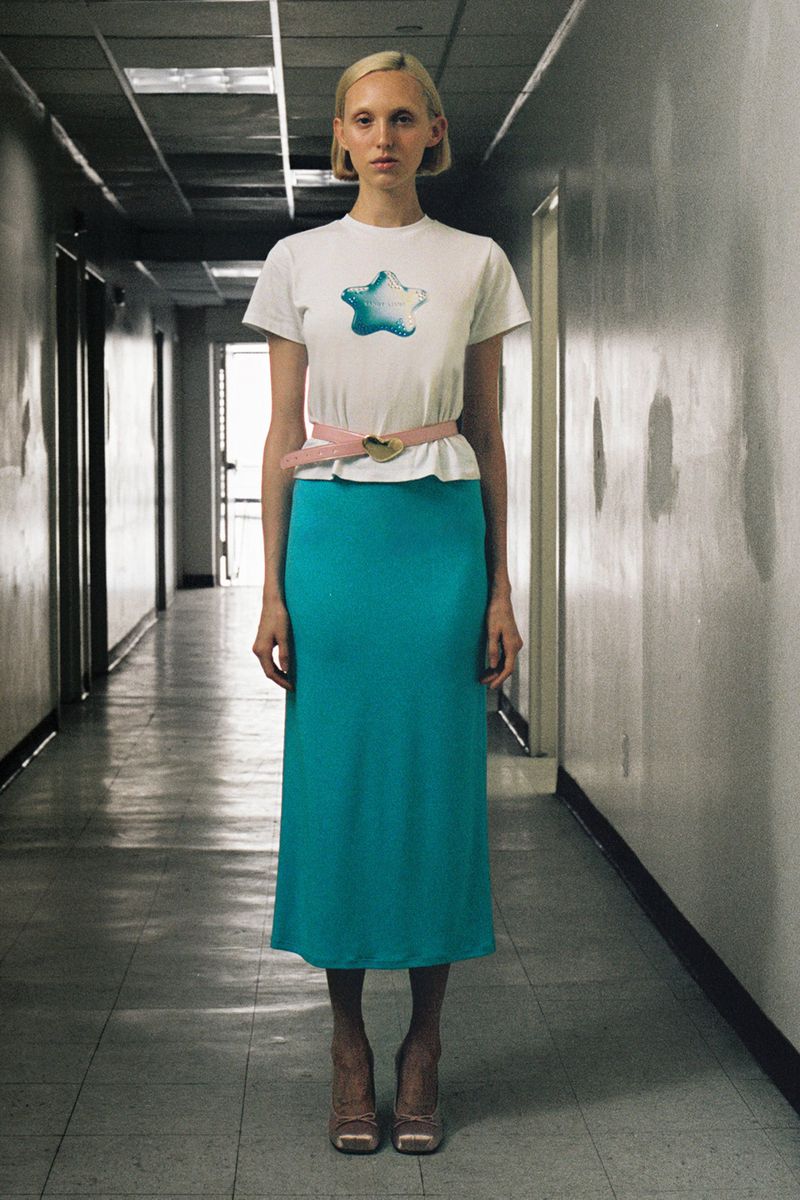 A woman in a white t-shirt, teal skirt, and pink belt stands in a tiled corridor with neutral lighting, facing the camera.