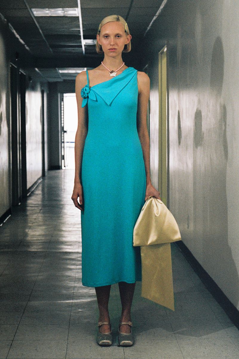A slender woman stands in a teal dress, holding a gold bag in a hallway. She has blonde hair and wears a pearl necklace.