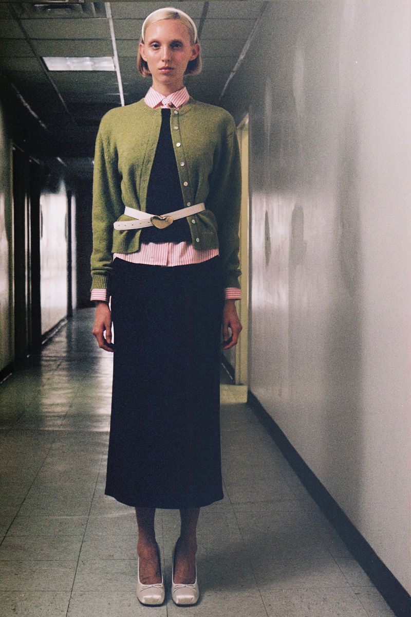A slender woman stands in a hallway wearing a light green cardigan, pink button-down shirt, long black skirt, and ivory high heels.