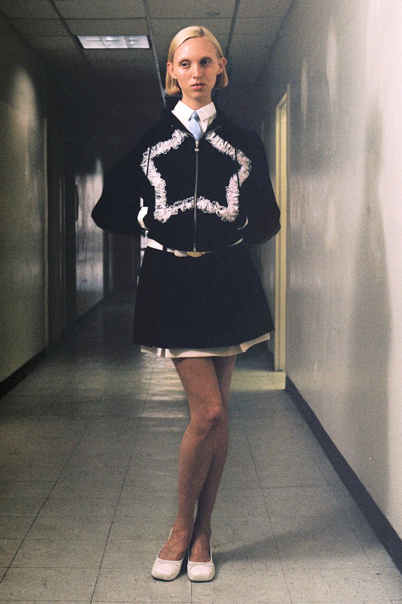 A full shot of a blond woman in a stylish black and white outfit and flats standing in a stark hallway with neutral colors.