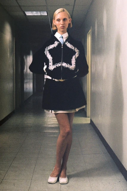A full shot of a blond woman in a stylish black and white outfit and flats standing in a stark hallway with neutral colors.