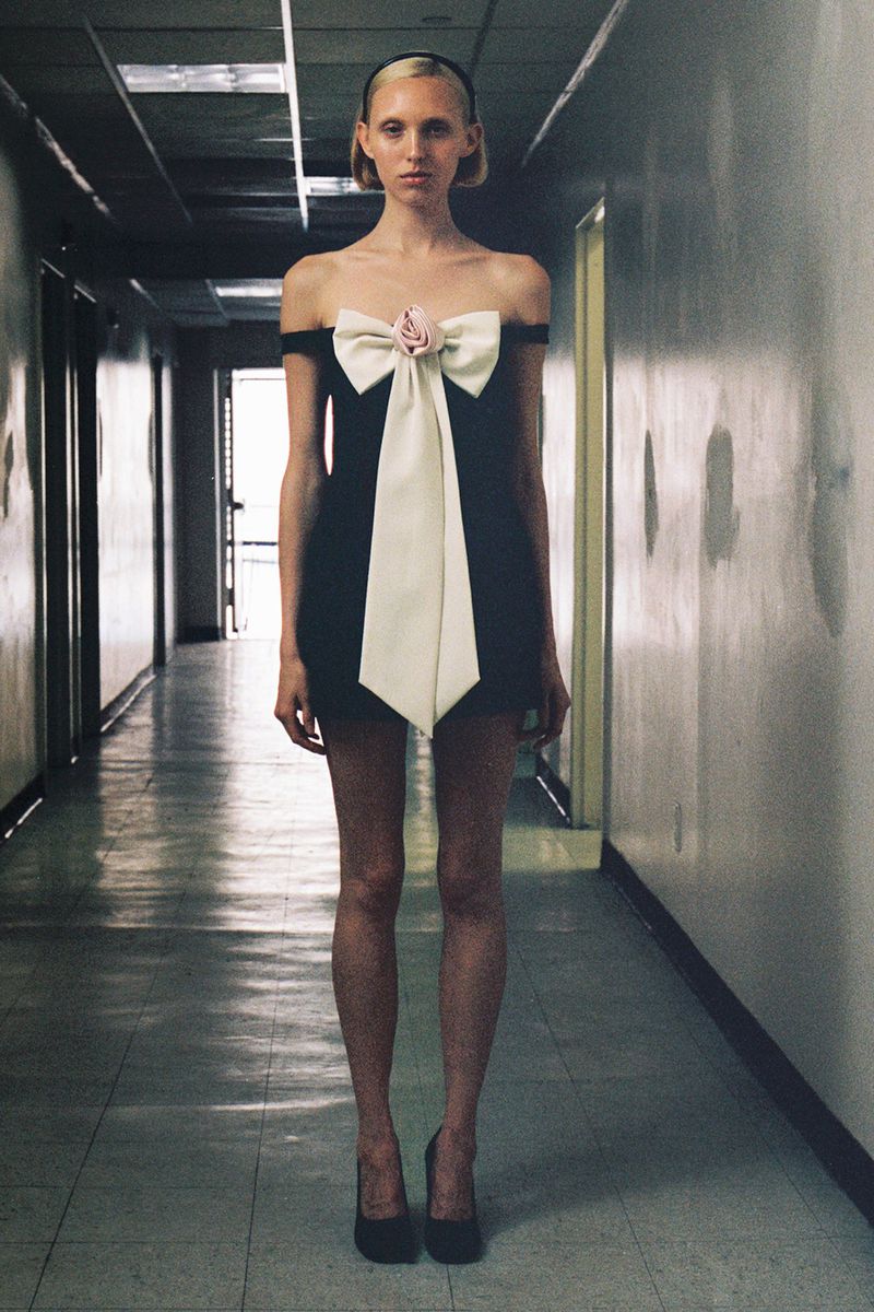 A slender adult female poses in a black dress with a large white bow in an indoor hallway, looking directly at the camera.