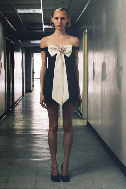 A slender adult female poses in a black dress with a large white bow in an indoor hallway, looking directly at the camera.