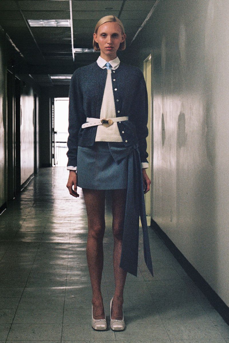 Fashion model poses in schoolgirl-style outfit in hallway with blue skirt, white shirt, and cardigan, illuminated by cool lighting.