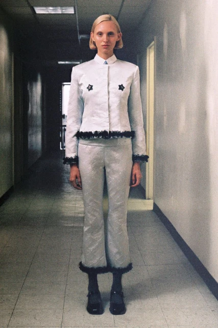 Adult female fashion model poses in a light gray suit with dark trim in an indoor hallway with a light gray floor and walls.