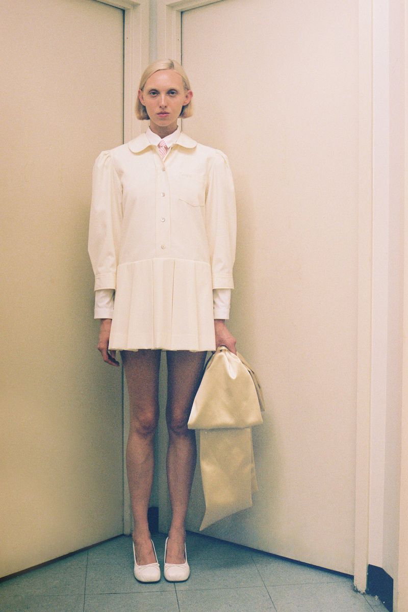 A slender woman with short blonde hair poses in an off-white dress and heels, holding a matching handbag in a corner setting.