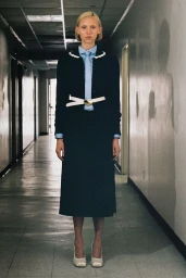 Model standing in a long hallway wearing a dark jacket, blue shirt, and white belt for Sandy Liang Resort 2025.