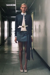 Model standing in a hallway wearing a dark cardigan, white collared shirt, skirt with sash, and belt for Sandy Liang Resort 2025.