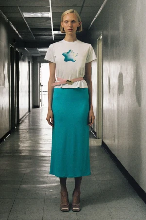 Model Delilah Koch in a white star graphic t-shirt, pink heart belt, and vibrant cyan midi skirt posing in a hallway.