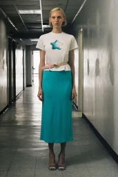 Model Delilah Koch in a white star graphic t-shirt, pink heart belt, and vibrant cyan midi skirt posing in a hallway.