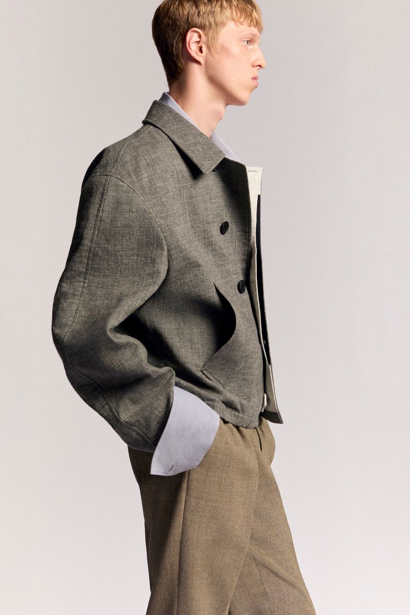 Profile shot of a blond male adult wearing a grey jacket over a light blue shirt with tan slacks in a studio setting.
