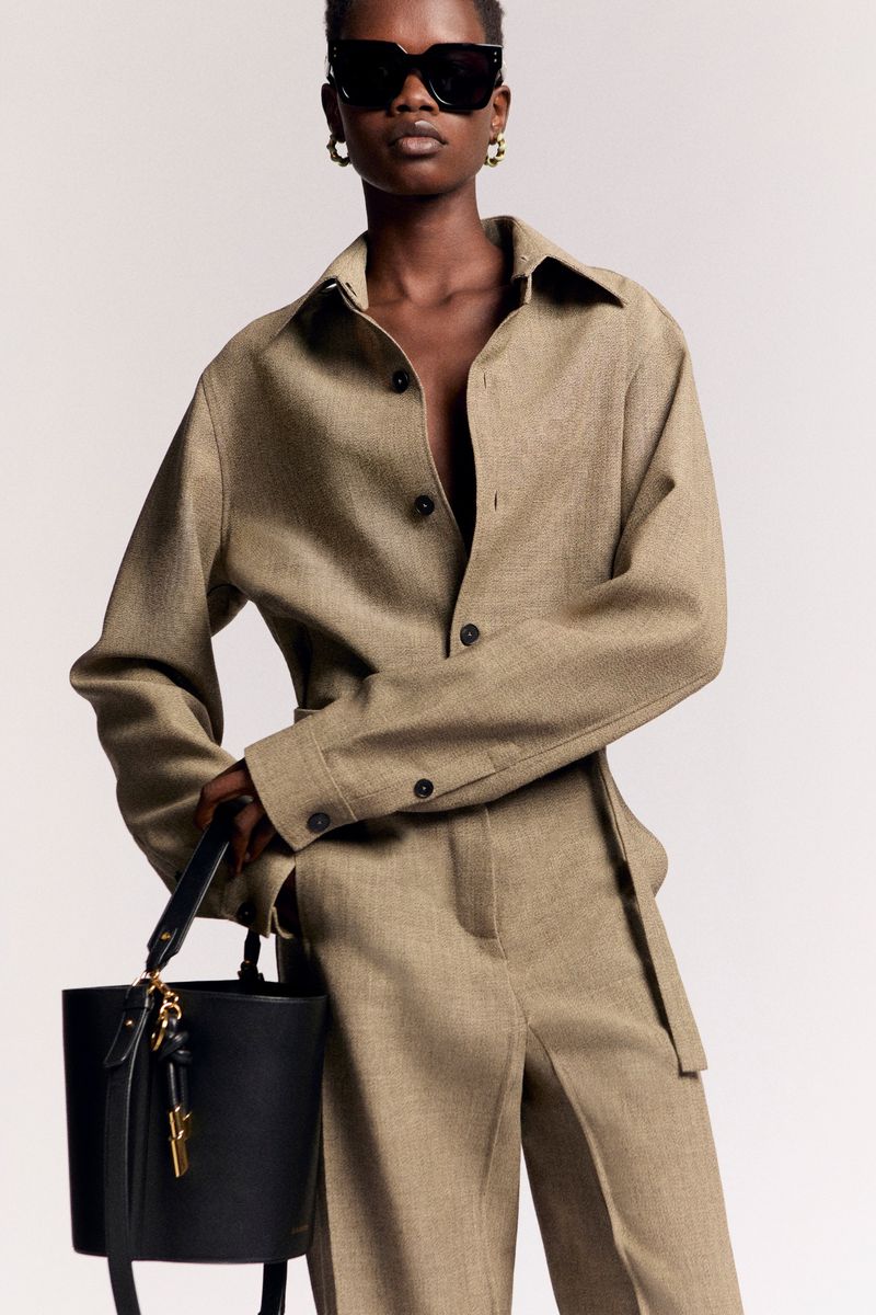 Fashionable adult female wearing a beige shirt and pants with a black bag and sunglasses in a studio setting with a white background.