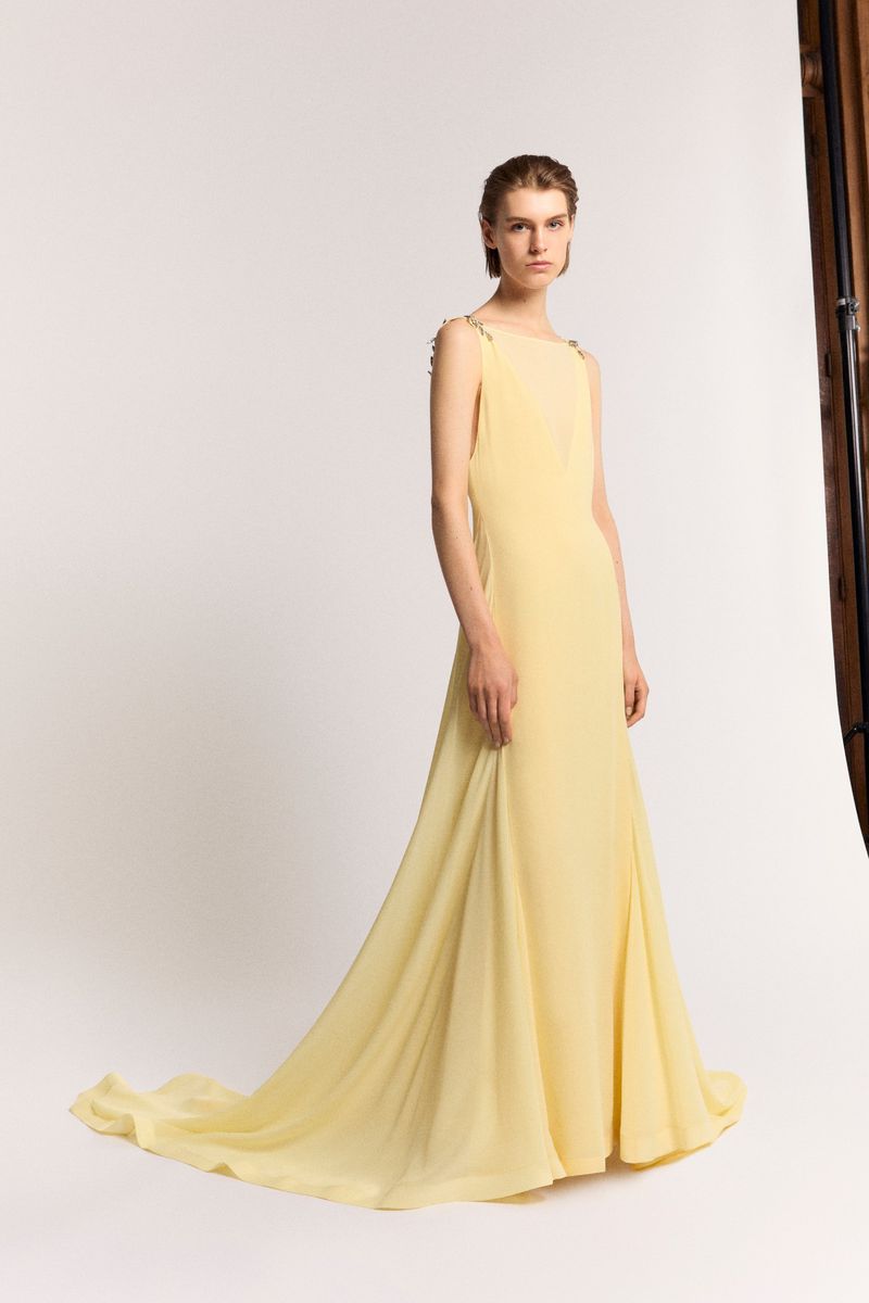 Elegant female model in a light yellow gown with a flowing train, posing in a photo studio with a minimalist backdrop. Fashion editorial.