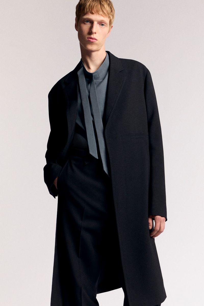 Portrait of a fair-skinned male model in a black coat and black trousers, posing in front of a clean, neutral-toned background.