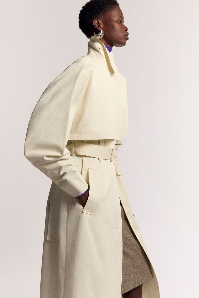 Elegant African-American woman poses in studio wearing a cream-colored trench coat with a lilac turtleneck sweater and statement earrings.