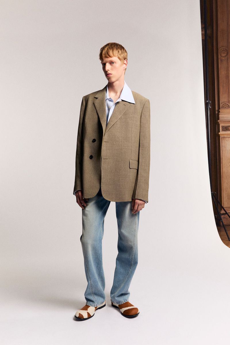 A fair-skinned male model poses in a light blue shirt, tan jacket, and light blue jeans in a photo studio setting with neutral lighting.