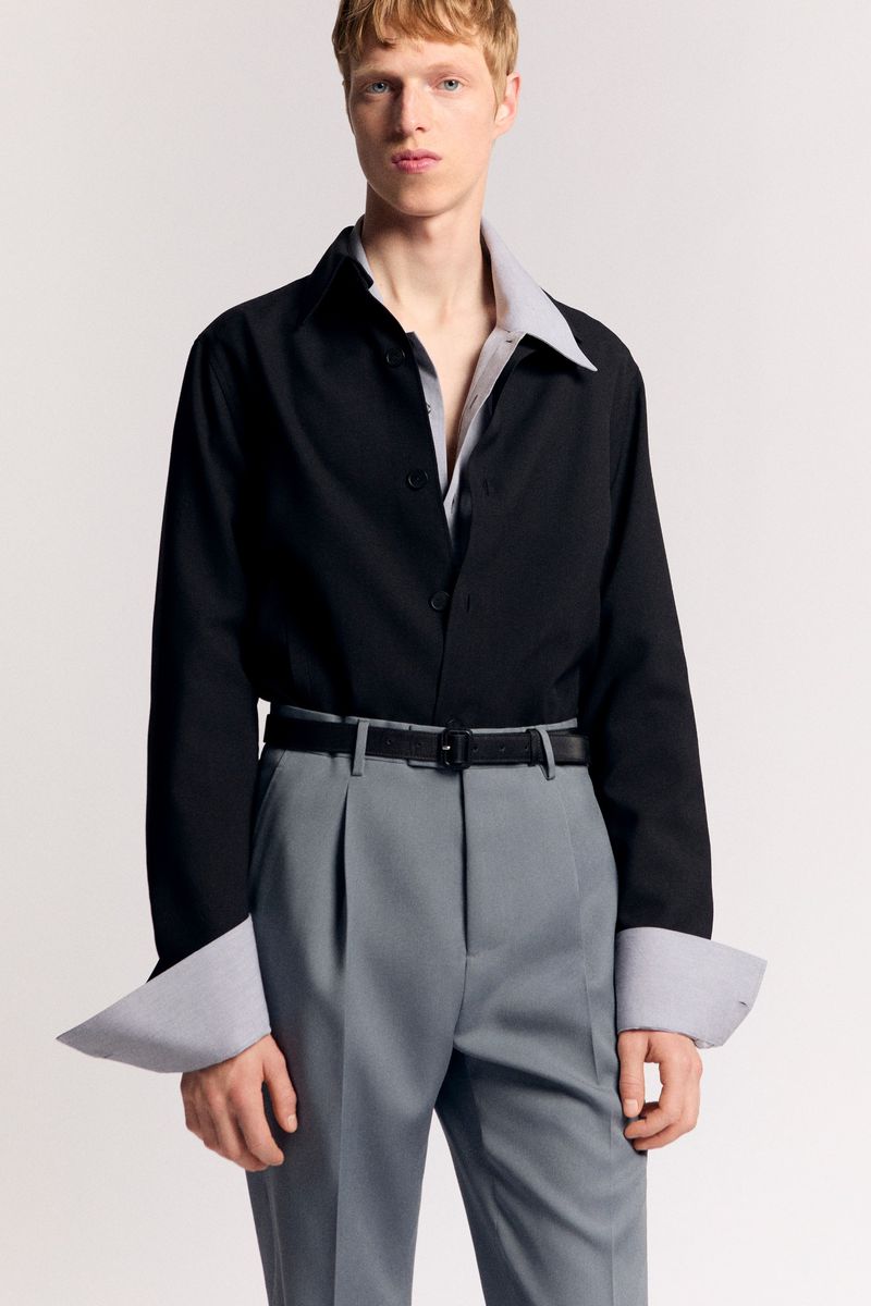 Fashion shot of a blond man in a black button-down shirt and grey pleated dress pants posing in a photo studio with a neutral backdrop.