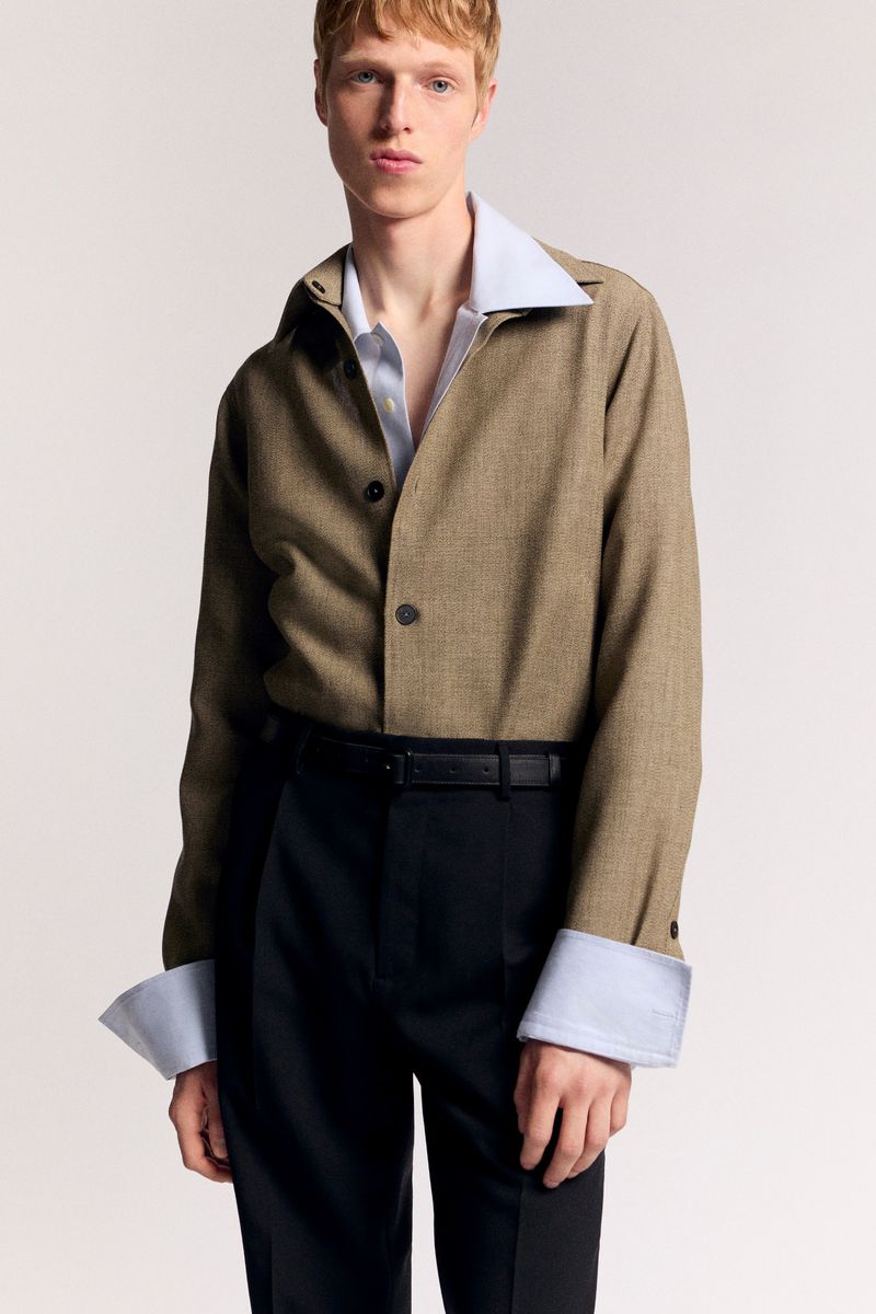 Model posing in a tan buttoned overshirt over a light blue collared shirt and black trousers against a pale seamless background.
