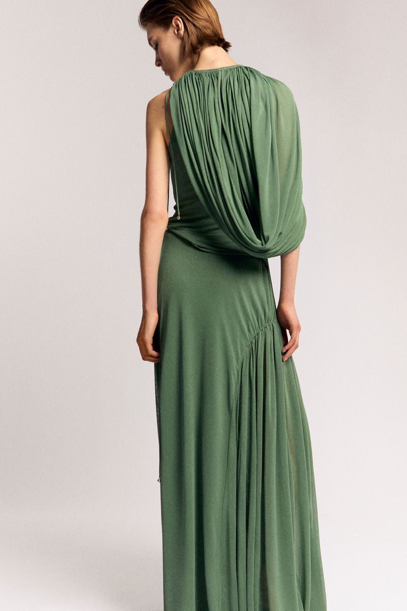 An adult female model poses in a long, draped, green dress against a light background in a studio setting, fashion photography.