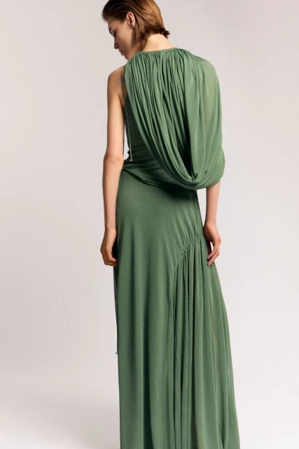 An adult female model poses in a long, draped, green dress against a light background in a studio setting, fashion photography.