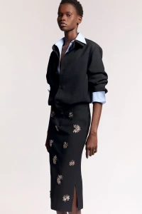 Model Mary Ukech wearing a black jacket over a pale blue collared shirt and an embellished black skirt against a white studio background.