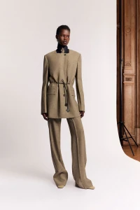 Model wearing a tan structured suit jacket and matching trousers against a white seamless studio backdrop.