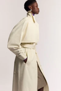 Model Mary Ukech in a belted cream trench coat and silver earring standing in profile against a neutral studio backdrop.