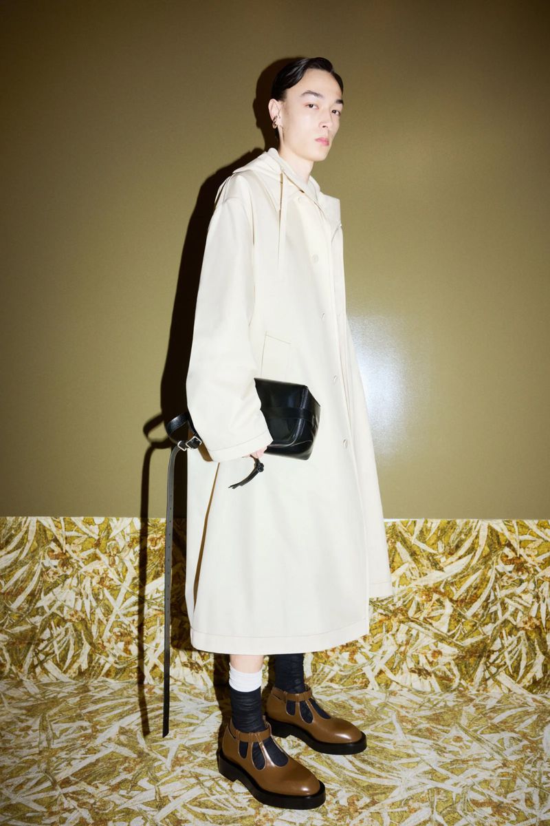 Young adult wearing cream-colored coat, black socks and brown shoes standing in a photo studio with olive-green wall and patterned floor.