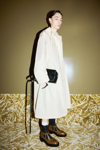 Young adult wearing cream-colored coat, black socks and brown shoes standing in a photo studio with olive-green wall and patterned floor.