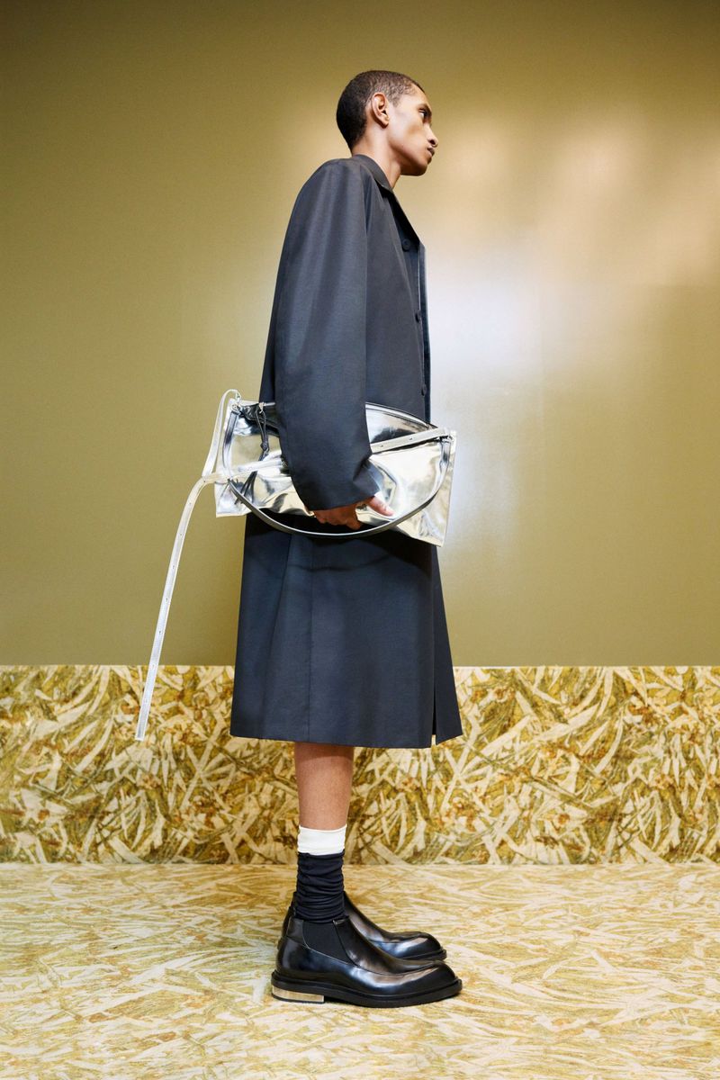 Fashion shot of a Black man in a black coat and skirt, carrying a silver bag, with black boots and white socks, against an olive wall.