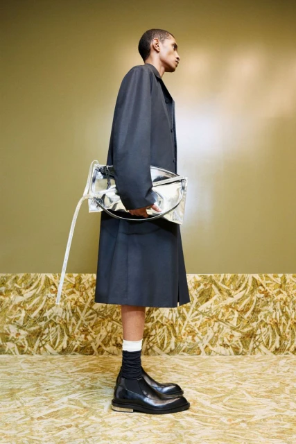 Fashion shot of a Black man in a black coat and skirt, carrying a silver bag, with black boots and white socks, against an olive wall.