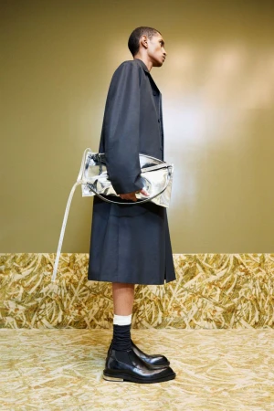 Fashion shot of a Black man in a black coat and skirt, carrying a silver bag, with black boots and white socks, against an olive wall.
